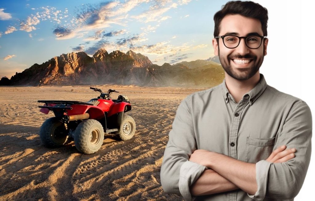 homme souriant les bras croisés et derrière lui il y a une photo d'un quad dans le désert.