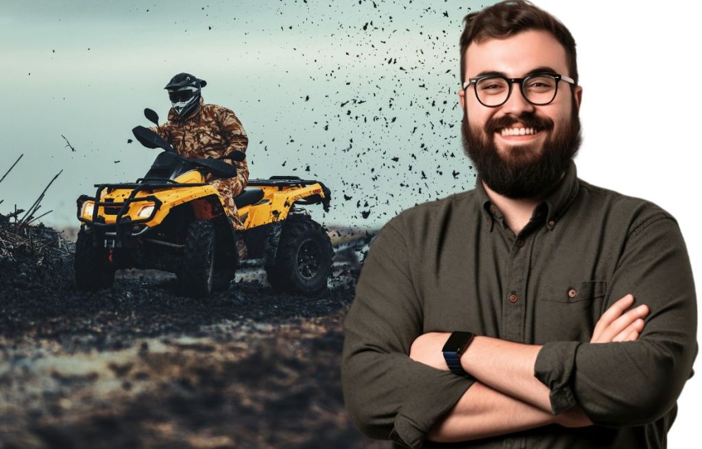 homme souriant les bras croisés. Derrière lui au second plan il y a un homme sur un quad qui roule dans la boue.