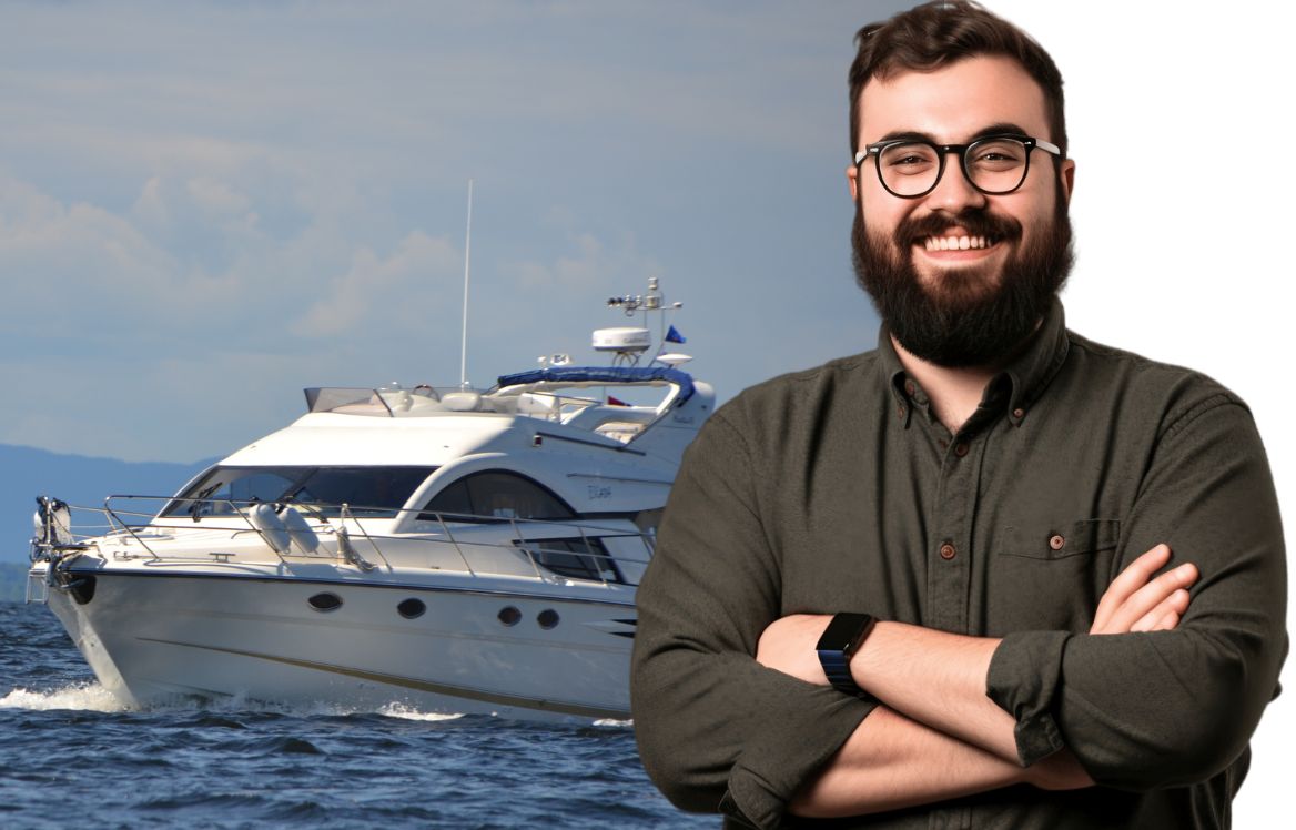 homme souriant à lunettes bras croisés au premier plan et un yacht en pleine mer derrière l'homme.