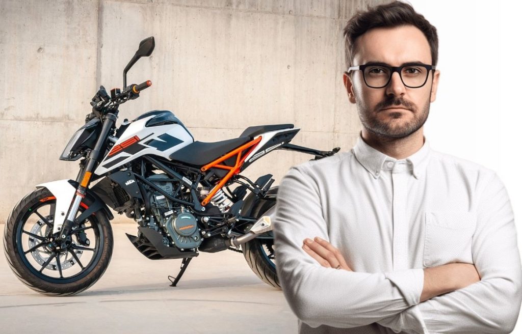 Homme sur de lui avec des lunettes qui vous regarde au premier plan et une moto orange blanche et noire au second plan qui est stationné vu de coté