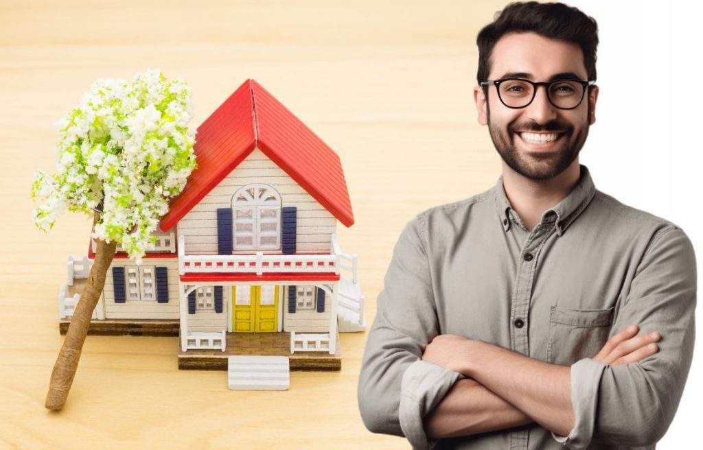 Homme souriant, les bras croisés et derrière lui il y à une maison type jouet posé sur un fond en bois