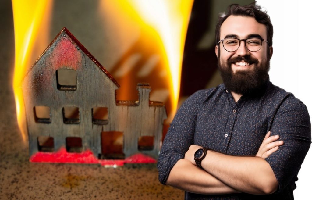 homme souriant bras croisés et en arrière plan il y à une maquette de maison en bois qui brûle.