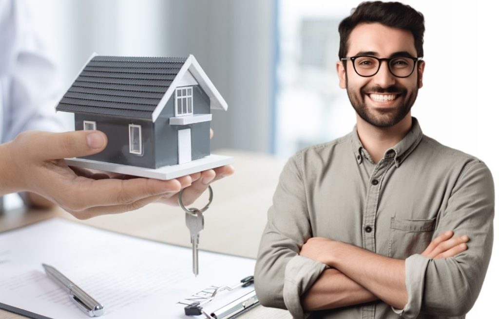 homme souriant et en arrière plan il y a une maison miniature qu'une mains nous tend avec des clés de maison