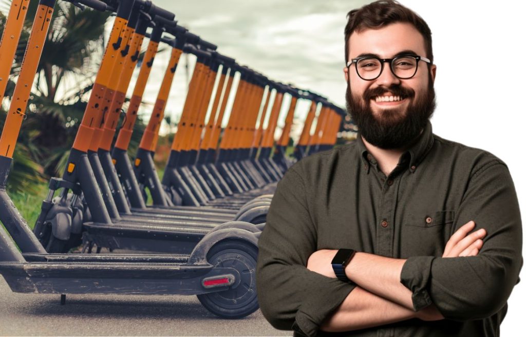 Un homme souriant les bras croisée au premier plan et derrière il y à de nombreuses trottinette électrique garé.