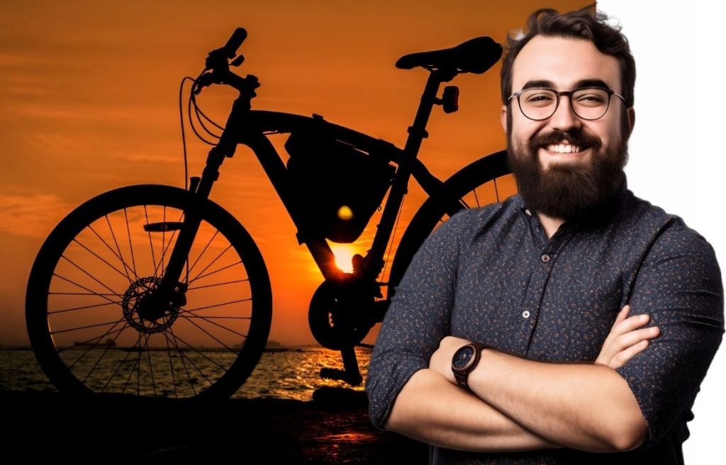 homme souriant, les bras croisés et en arrière plan il y à un vélo devant un couché de soleil