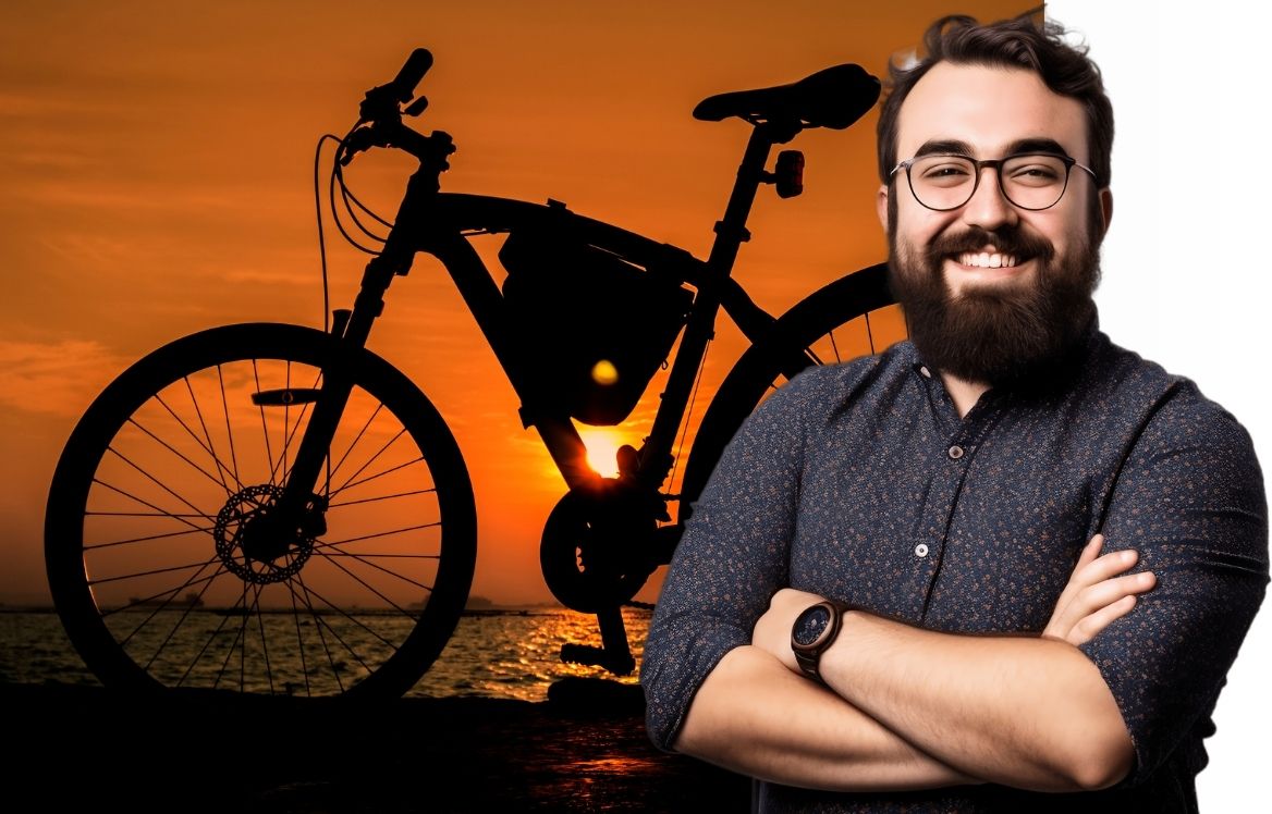 homme souriant, les bras croisés et en arrière plan il y à un vélo devant un couché de soleil